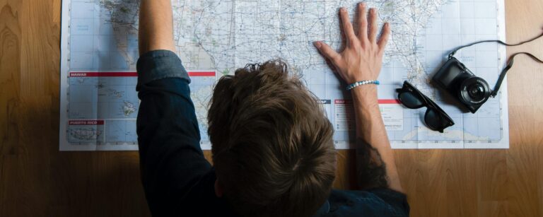 Top view of anonymous young male in casual clothes sitting on wooden floor with photo camera and coffee cup and pointing on paper map while planning trip route